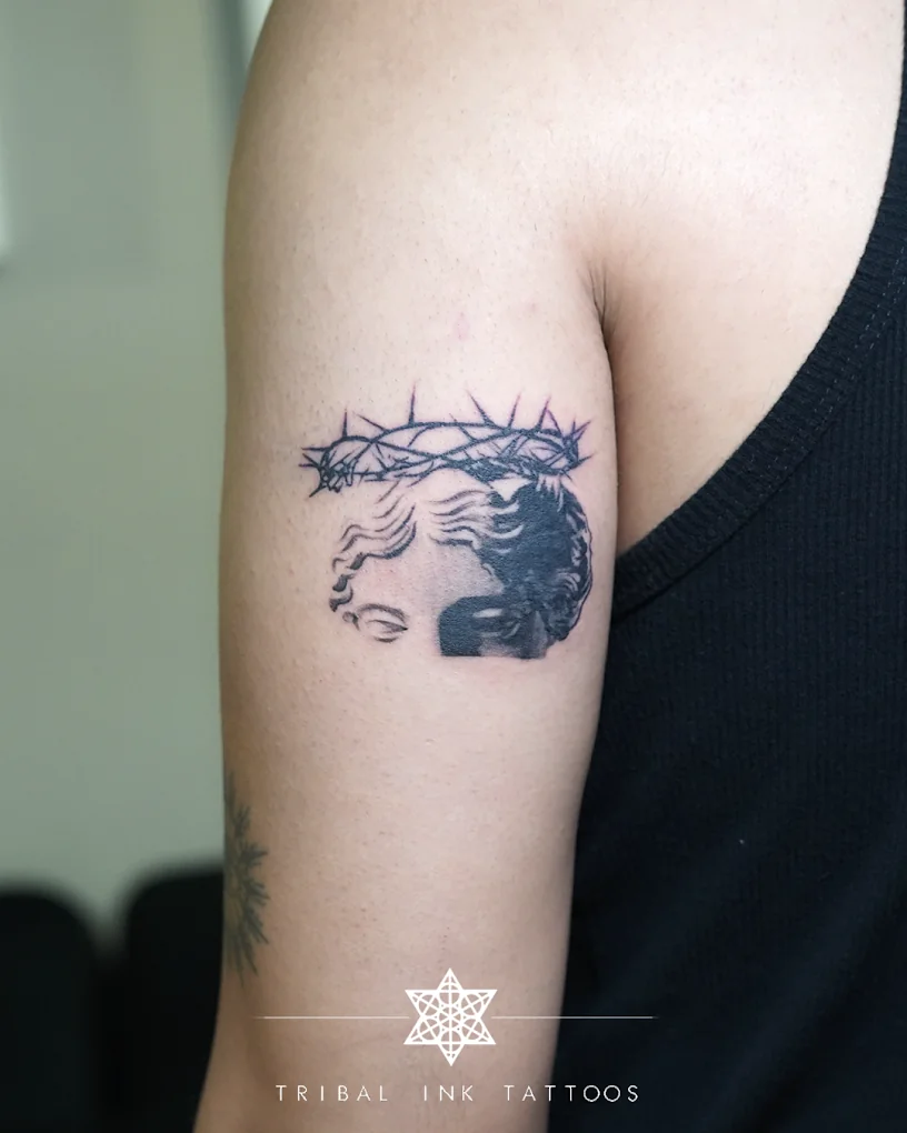 A classical head wearing a crown of thorns, tattooed small in micro-realism on an upper arm.