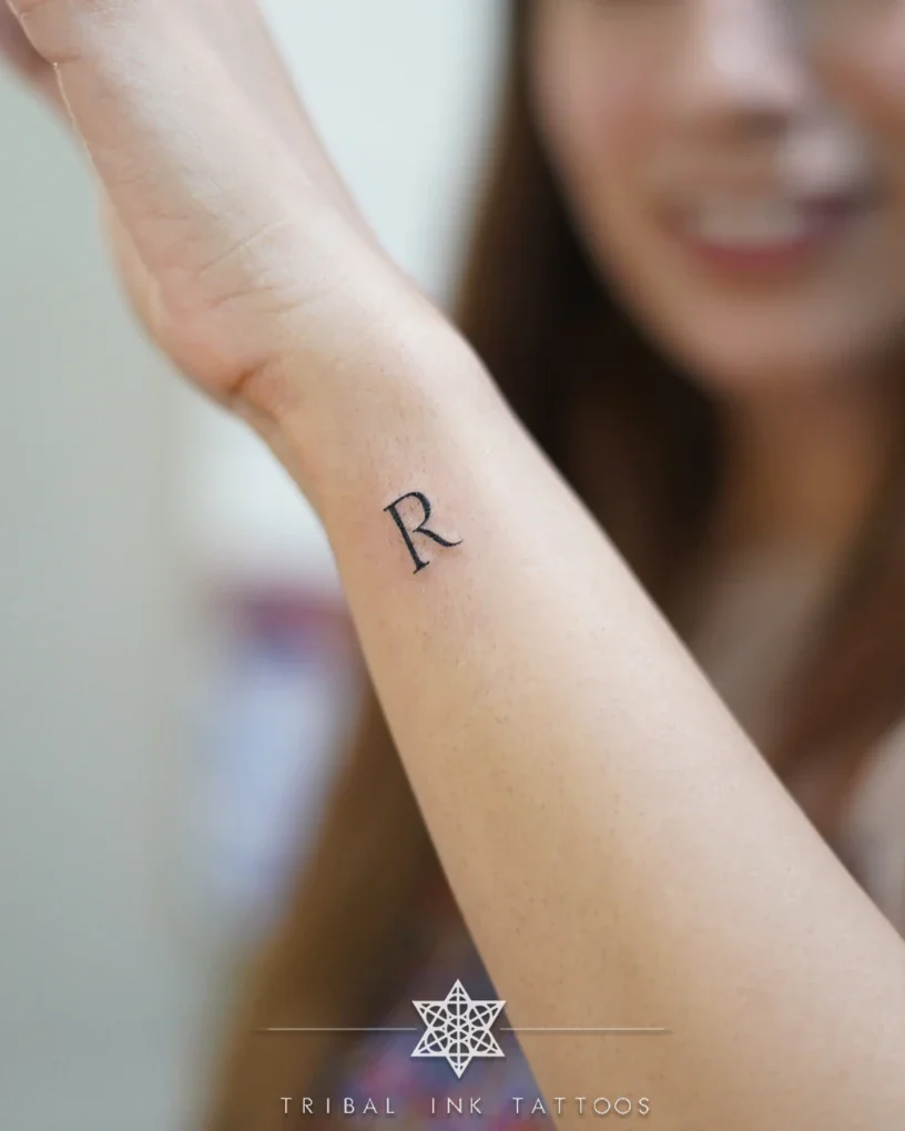 A single serif letter R tattooed in fine line on the inside of a woman's wrist.