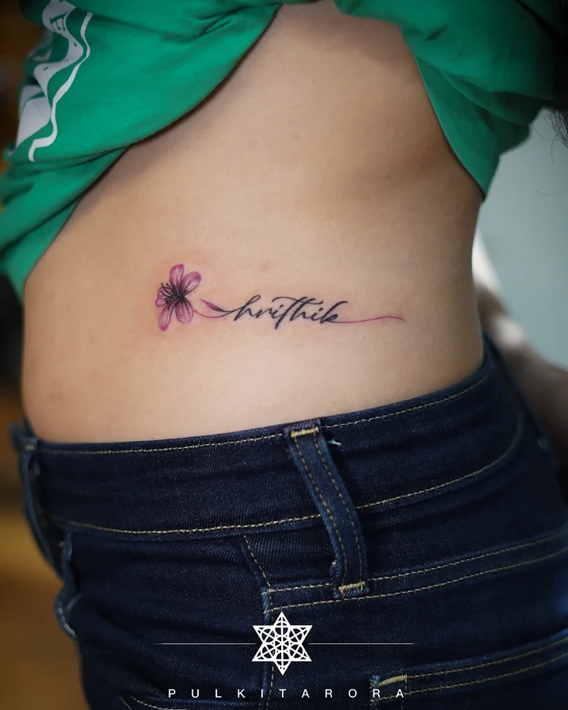 A name written in fine script with a small pink flower, tattooed on a woman's waist.