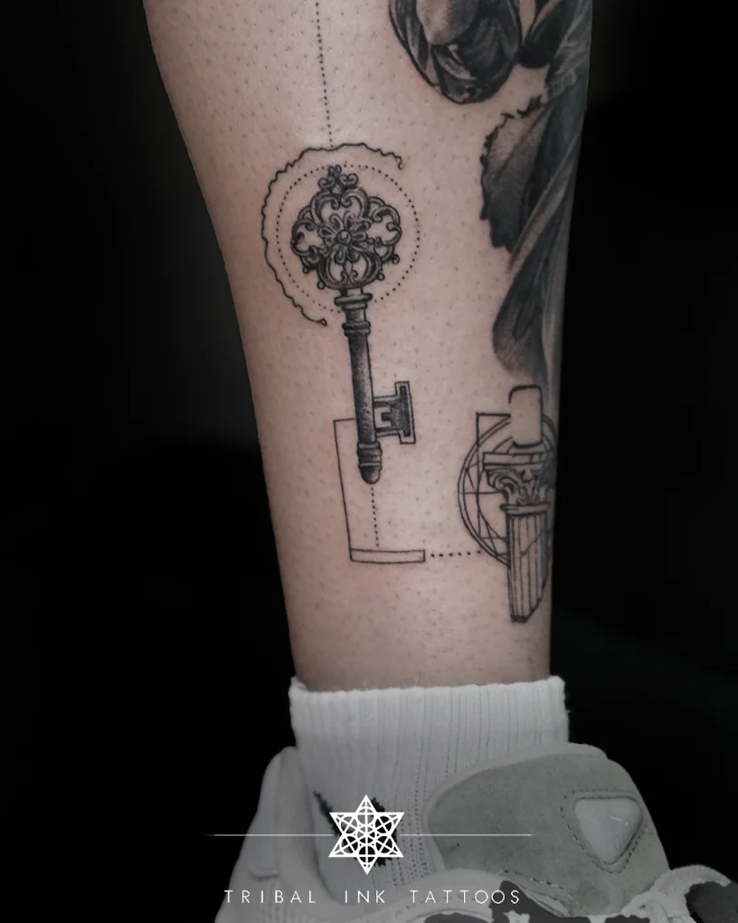 An ornate key with fine geometric linework and a dotted circle, tattooed on a calf.