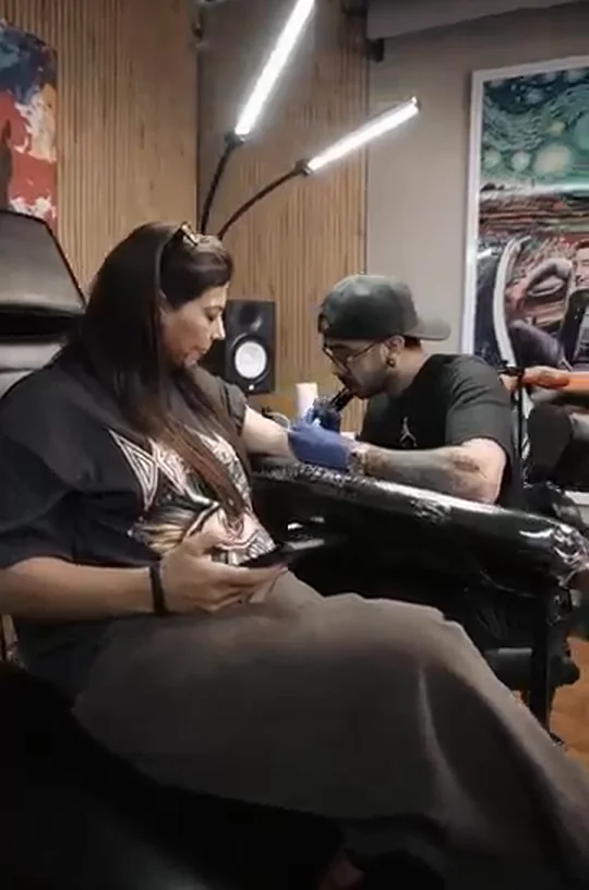 An artist working on a client's arm at Tribal Ink Tattoos, under a strip light against a wooden slat wall.