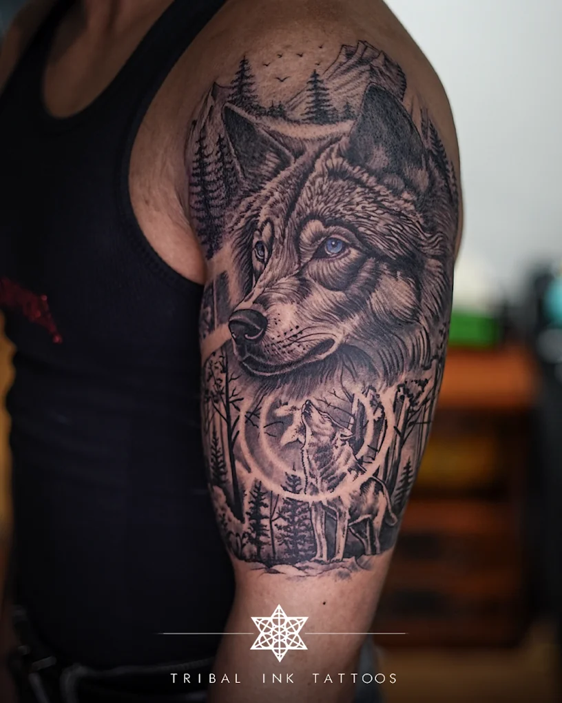 A wolf's face above a pine forest and a howling wolf, tattooed in black and grey across a man's upper arm.